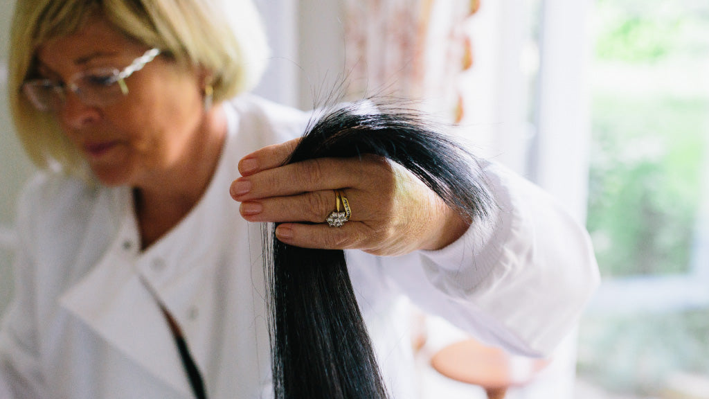 Photo showing Eva Proudman, a white female trichologist, wearing a white lab coat and inspecting a person's long dark hair